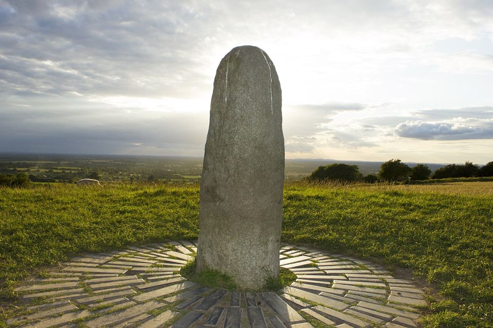Pierre dressée au sommet d'une colline herbeuse, entourée d'un pavage rayonnant, sous un ciel nuageux